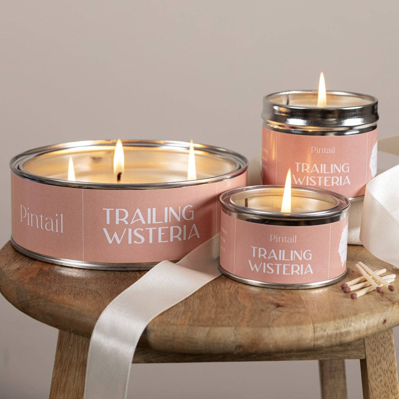 Three 'Trailing Wisteria' candles in pink tins on a wooden surface with a neutral background.