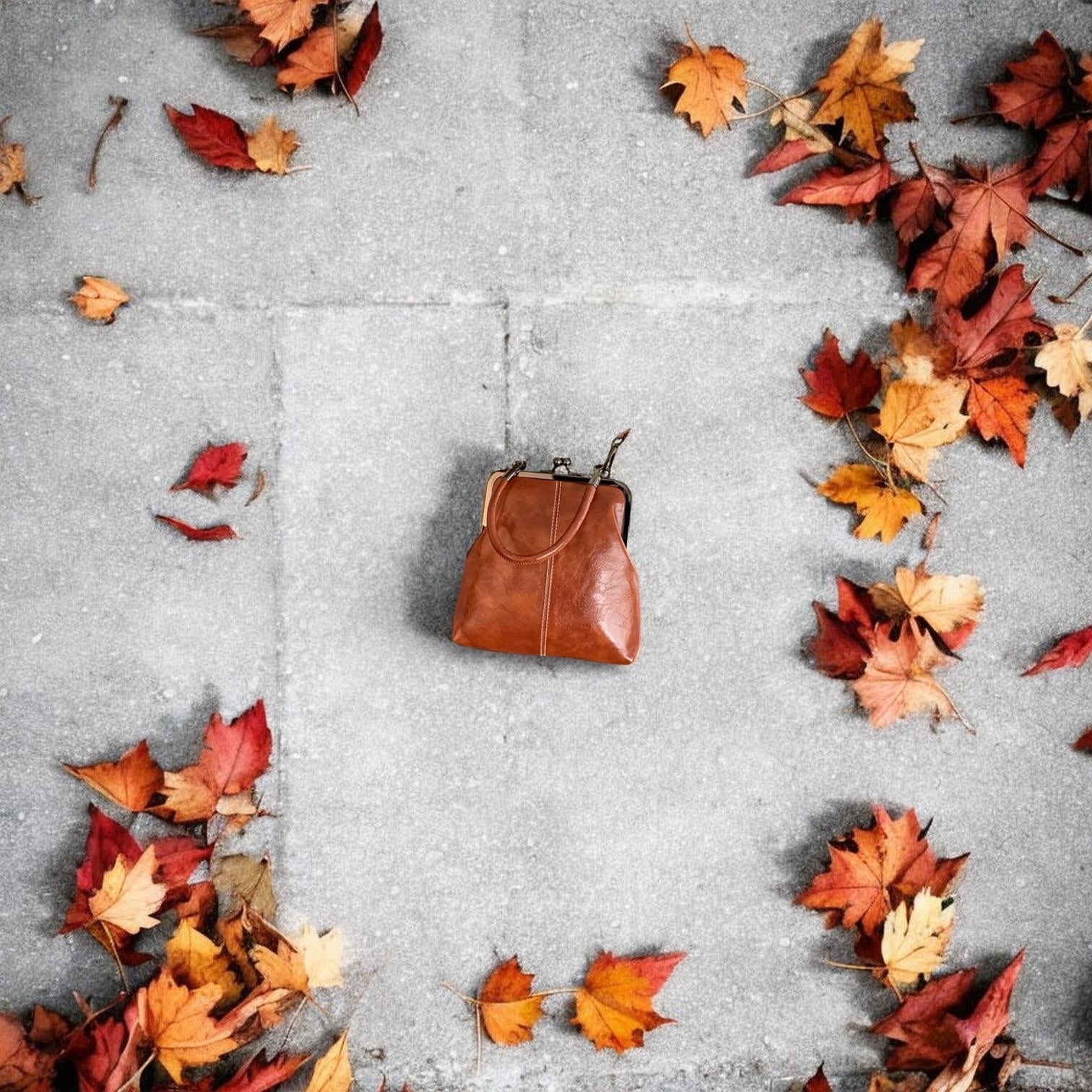 Brown leather handbag on a gray concrete surface with autumn leaves scattered around.