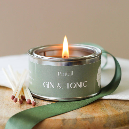 Candle in a tin labeled 'Pintail Gin & Tonic' on a wooden surface with a green ribbon.