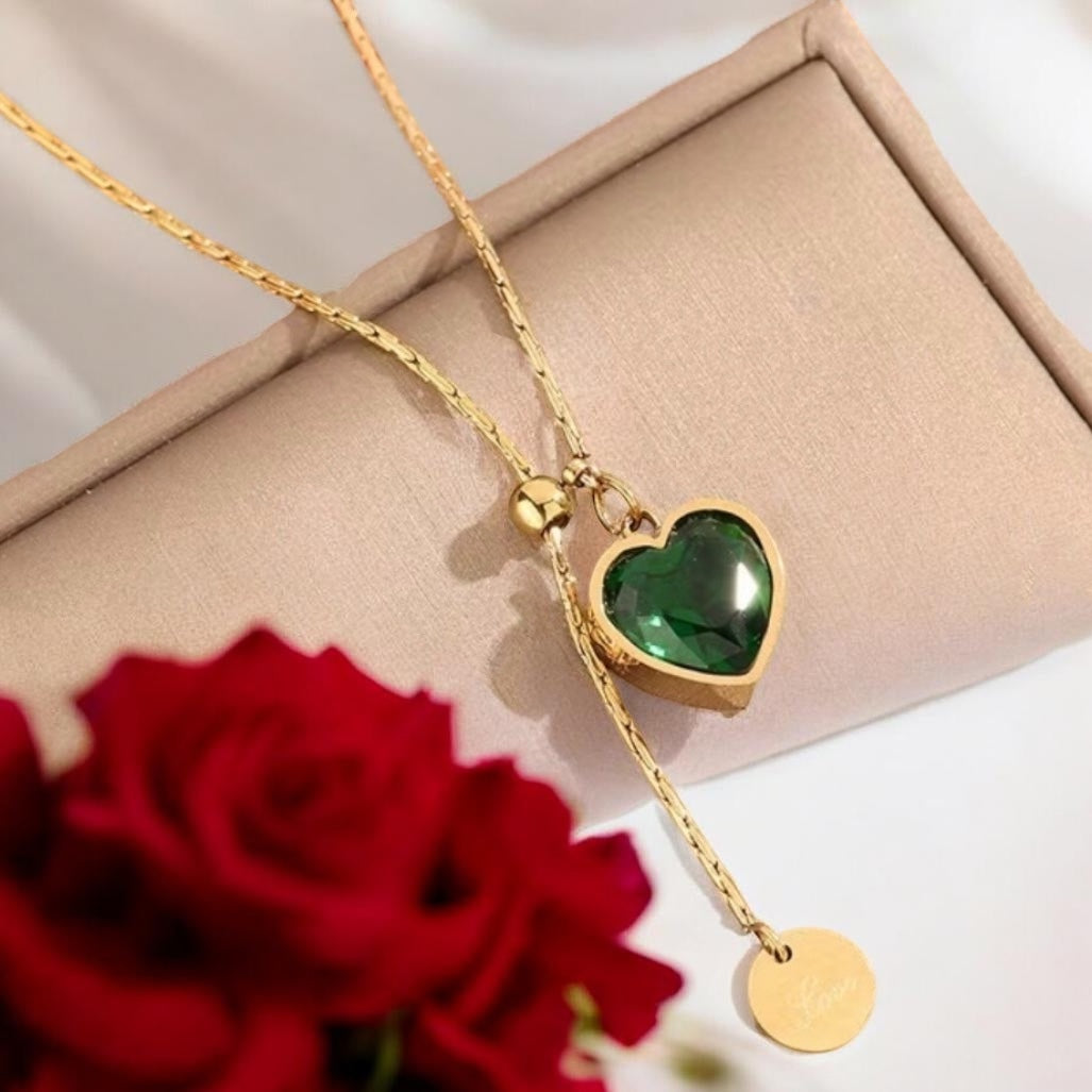 Gold necklace with a green heart pendant on a beige surface with a red rose.