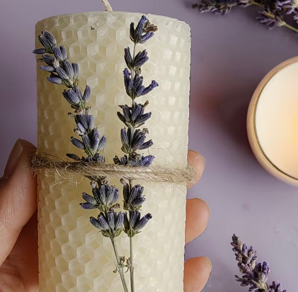 Hand holding a candle with lavender flowers wrapped around it on a soft purple background.