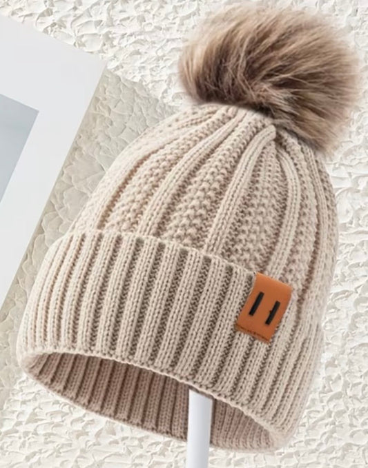 Beige knit beanie with a fluffy pom-pom on a textured surface