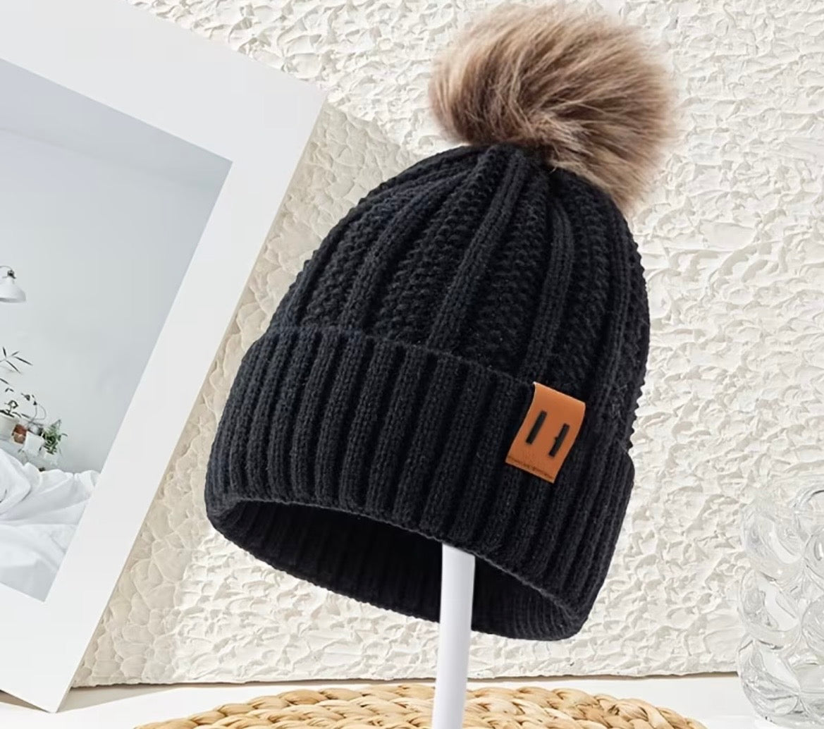 Black knit beanie with a brown pom-pom on a white stand against a textured white background.