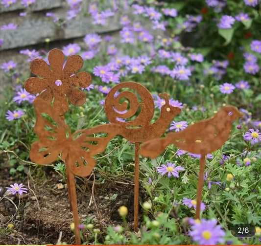 Decorative metal garden stakes shaped like a butterfly, flower, and bird in a garden with purple flowers.