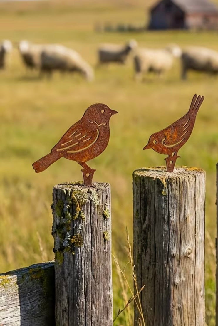 Two rusted metal bird sculptures on wooden posts with sheep grazing in the background.