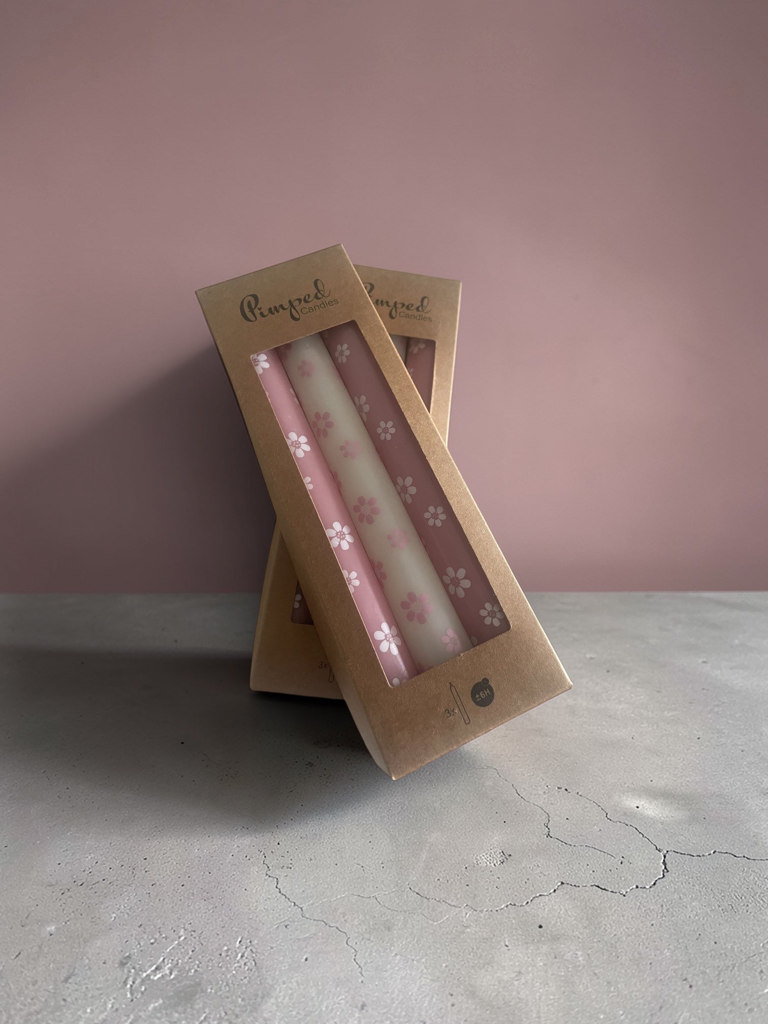 Two pink candles with floral design in a cardboard packaging against a pink background