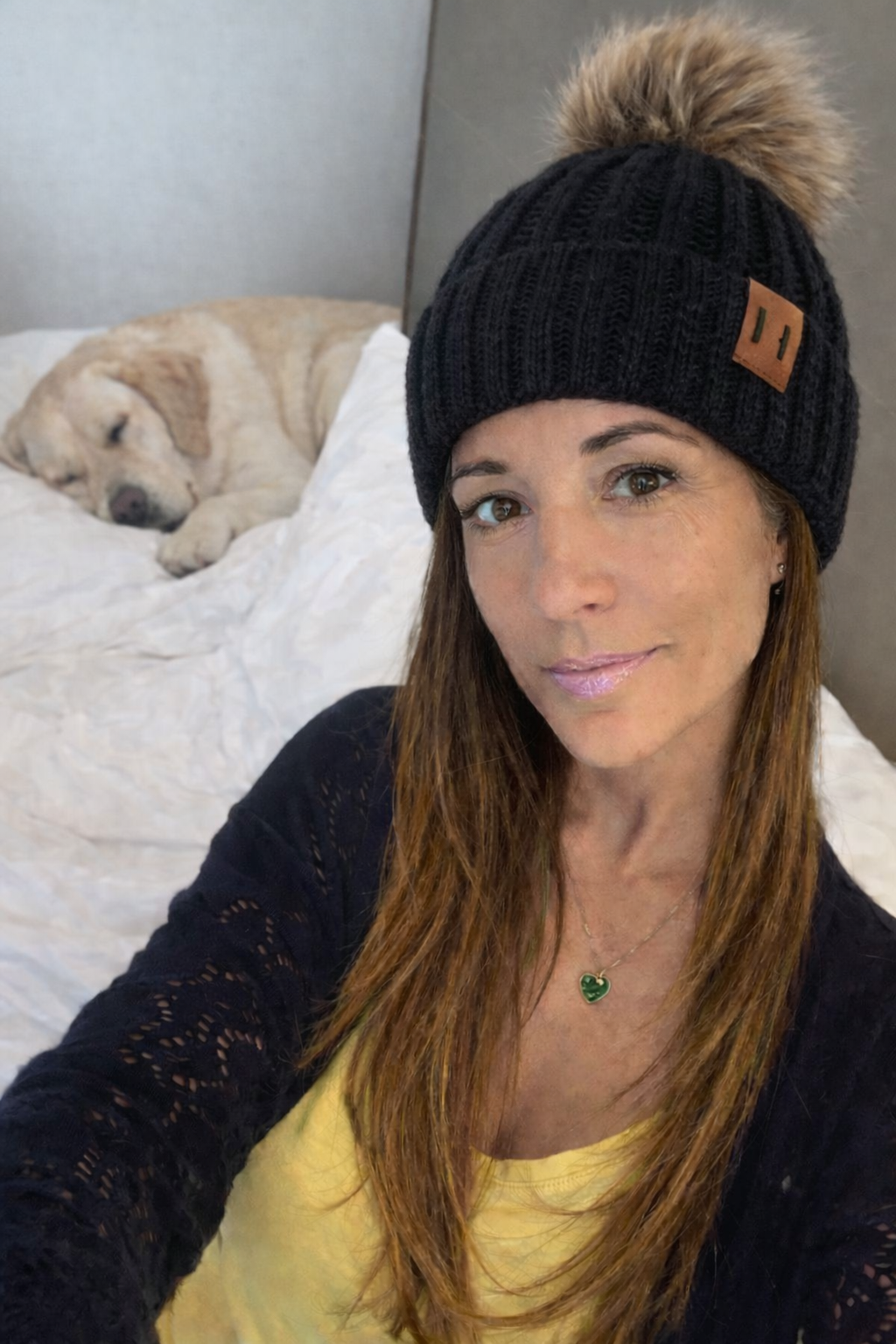 Woman wearing a black beanie with a pom-pom, sitting on a bed with a dog in the background.