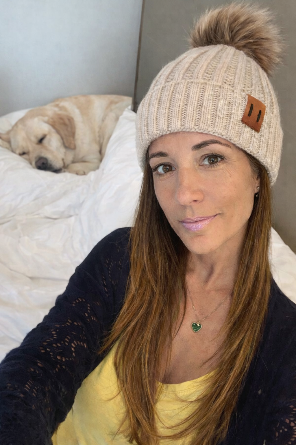 Woman wearing a beige knit beanie with a pom-pom, sitting on a bed with a dog in the background.