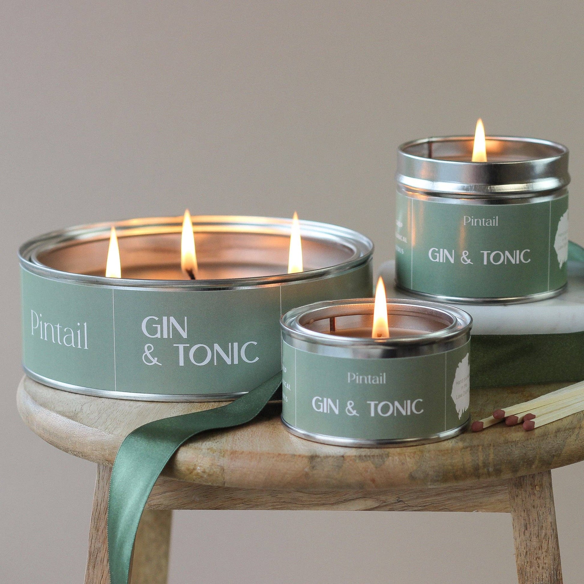 Three 'Gin & Tonic' candles in metal tins on a wooden stand with a plain background.